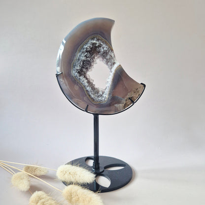 Handgeslepen agaat maan op zwarte metalen standaard met natuurlijke druzy geode kristallen en zachte grijsbruine banding.