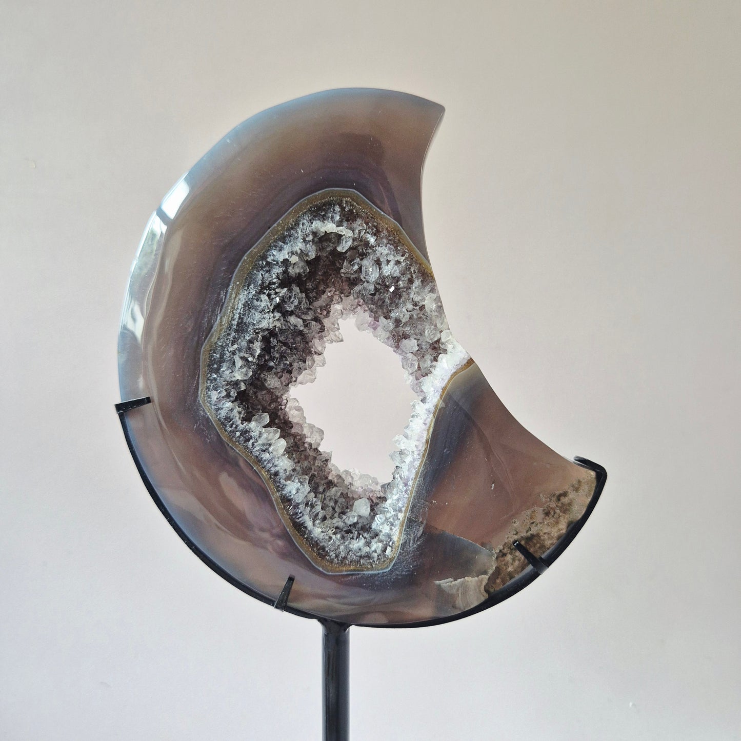 Handgeslepen agaat maan op zwarte metalen standaard met natuurlijke druzy geode kristallen en zachte grijsbruine banding.